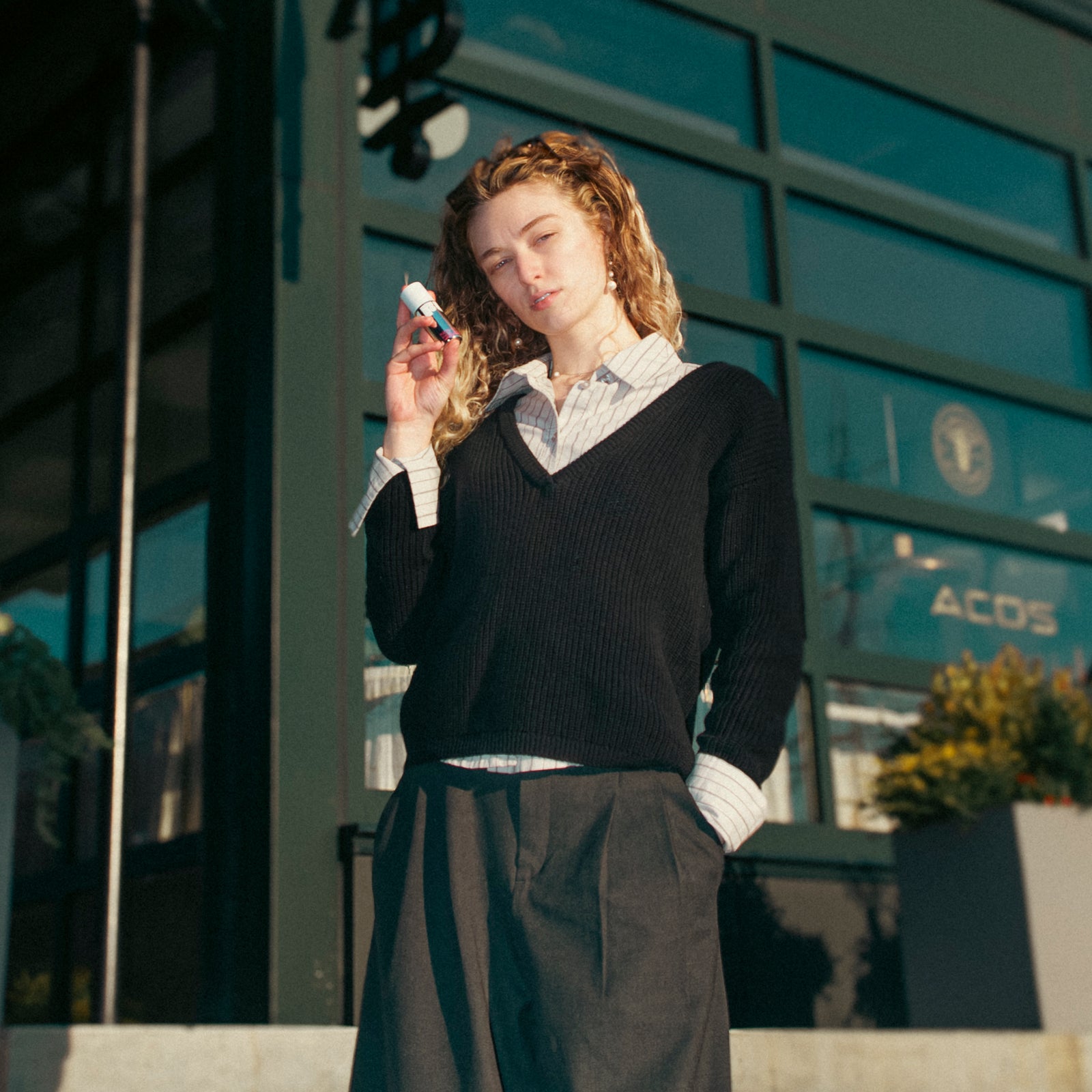 Woman standing outdoors holding a phone, wearing a black sweater and dark pants.